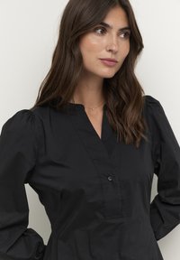 Haut noir à boutons avec un col en V, manches bouffantes longues et texture de tissu lisse. Design minimaliste et silhouette ajustée.