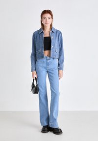Jeune femme portant une veste courte bleue, un top bandeau noir, un jean taille haute bleu clair, des bottes noires, tenant un petit sac à main noir, debout sur un sol gris.