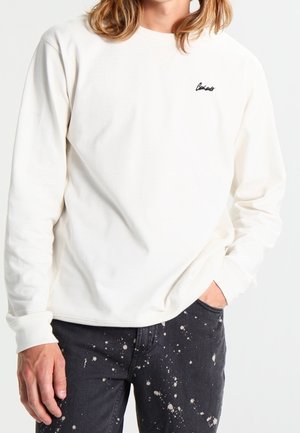 Osoba ubrana w białą koszulę z długim rękawem z małym czarnym napisem na piersi oraz czarne jeansy z białymi plamami farby.