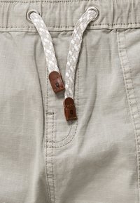 Shorts beige clair avec une taille élastique, dotés d'un cordon tissé crème et blanc et d'accents en cuir marron. Tissu texturé avec des rayures subtiles.