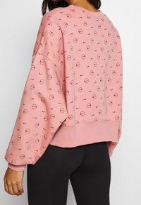 Rosa sweatshirt med ett upprepat mönster av små cirklar och Nike-logotyper. Den har en kortad design med vida ärmar och en mjuk textur.