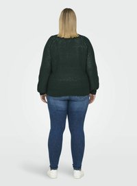 Pulover tricotat de culoare verde închis, cu o croială lejeră, mâneci ribelate și un gât larg, asortat cu blugi strâmți albaștri și adidași albi.