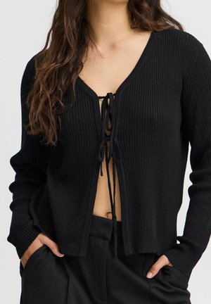 Femme portant un cardigan en maille côtelée noire noué avec deux cordons à l’avant, associé à un pantalon noir, mains dans les poches.