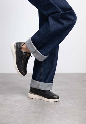 Zapatillas de cuero negro con suelas de goma blanca, con cordones y una cremallera lateral, combinadas con pantalones vaqueros oscuros de mezclilla azul con puños.