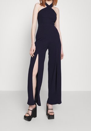 Granatowy kombinezon z dekoltem halter, z bocznymi rozcięciami w szerokich nogawkach. Modelka ma na sobie czarne sandały na platformie.