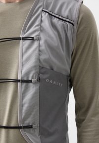 Lätt grå väst med nätpaneler, elastiska snören och zip-fickor. Har Oakley-logotyp i en subtil, präglad design.