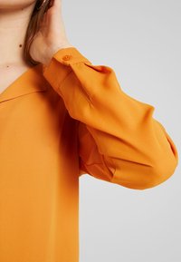 Blusa arancione con una texture morbida e fluida. Presenta una vestibilità rilassata e dettagli con polsini abbottonati. Design semplice con colletto sciallato.