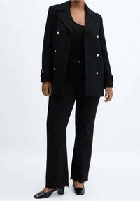 Blazer negro cruzado con botones dorados, combinado con pantalones negros con dobladillo sin rematar. Zapatos negros brillantes de punta afilada.