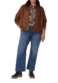 Frau in braunem Wildlederjacke, gemusterter Bluse, blauen weiten Jeans und schwarzen Plateaustiefeln steht mit Händen in den Taschen.