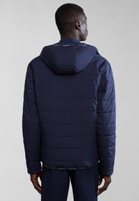 Giacca puffer blu scuro con design trapuntato, cappuccio e polsini a costine. Texture liscia con orlo dritto e dettagli blu lungo il colletto.