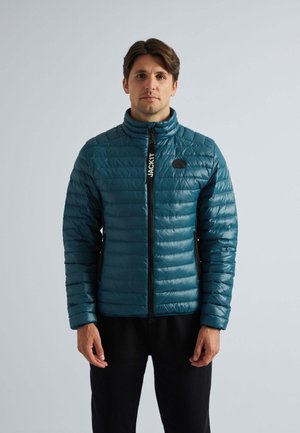 Giacca puffer teal con texture trapuntata, colletto alto, chiusura frontale con zip e dettagli neri. La modella la indossa sopra un outfit nero.