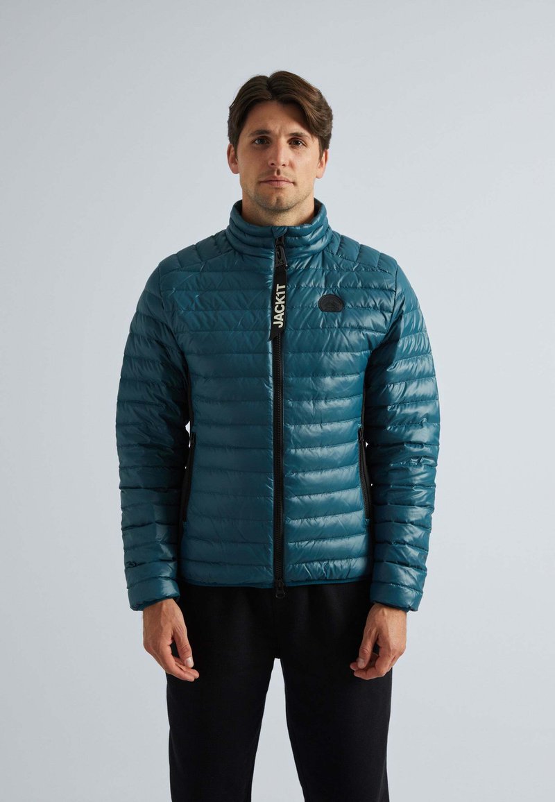 Veste en duvet teal avec texture matelassée, col haut, fermeture à glissière sur le devant et accents noirs. Le mannequin la porte par-dessus une tenue noire.