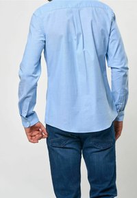 Man i ljusblå långärmad skjorta med knappar och mörkblå jeans, sedd bakifrån mot en enfärgad bakgrund.