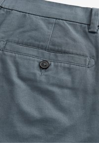 Gros plan sur un pantalon en tissu bleu-gris montrant une poche arrière avec un bouton noir et un détail de passant de ceinture.