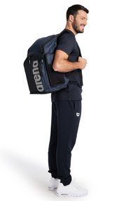 Sac à dos bleu et noir avec un devant en mesh, détail de logo et plusieurs poches zippées. Le modèle porte un pantalon bleu marine et des baskets blanches.