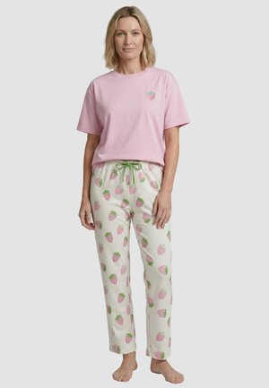 Vrouw draagt een lichtroze korte mouwen shirt en witte pyjamabroek met roze aardbeienprint en een groene trekkoord, staat op blote voeten.