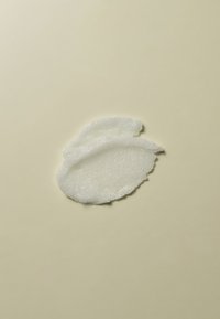 Gommage exfoliant blanc avec une texture crémeuse et épaisse et des granulés visibles, étalé sous une forme organique et arrondie sur un fond beige clair.