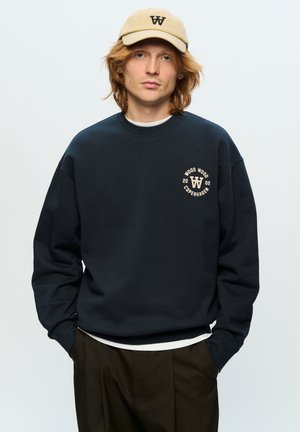 Dark navy sweatshirt with round neckline and elastic cuffs, featuring a beige circle logo on the chest. Paired with a beige cap.