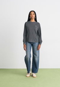 Chemise grise à manches longues avec un petit logo, associée à un jean bleu large et des baskets blanches, se tenant sur un sol vert clair.