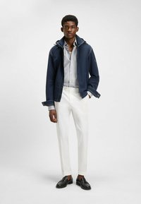 Mörkblå zip-up jacka med huva och vit sömnad, kombinerad med en ljusrandig skjorta och vita smala byxor. Svarta loafers fulländar looken.