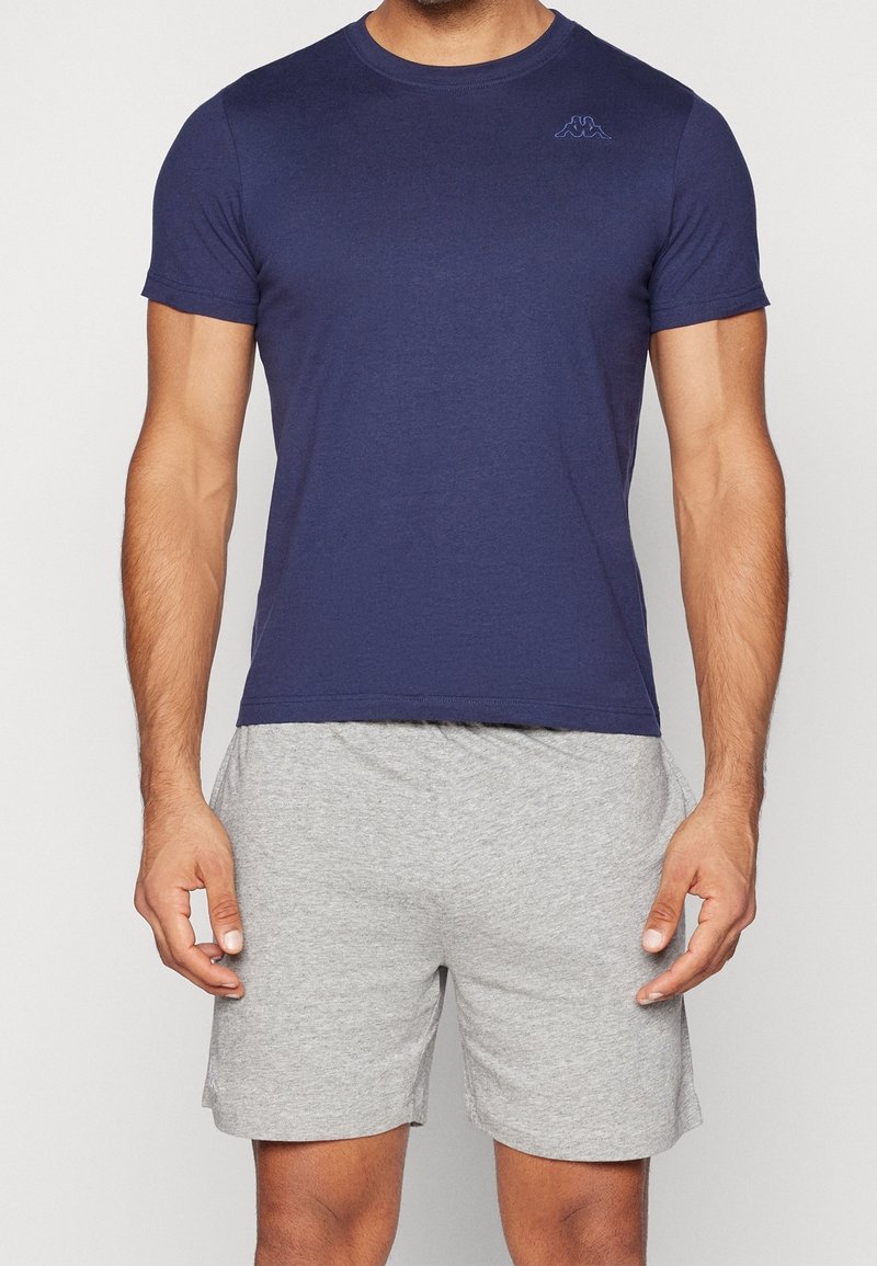 Homme portant un t-shirt bleu marine à manches courtes et un short de sport gris clair, debout devant un fond uni.