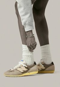 Beige und braune Wildledersneaker mit weißen Schnürsenkeln, strukturierten Akzenten und gelben Sohlen, kombiniert mit weißen gerippten Socken und grauen Jogginghosen.