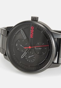 Reloj analógico negro con tres subesferas, que presenta una esfera negra lisa, acentos rojos y una correa de acero inoxidable. Marcaciones para los días y las horas.