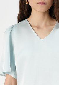 Top azzurro chiaro con scollatura a V e maniche corte a sbuffo. Tessuto morbido con una leggera lucentezza. Design minimalista e linee pulite.
