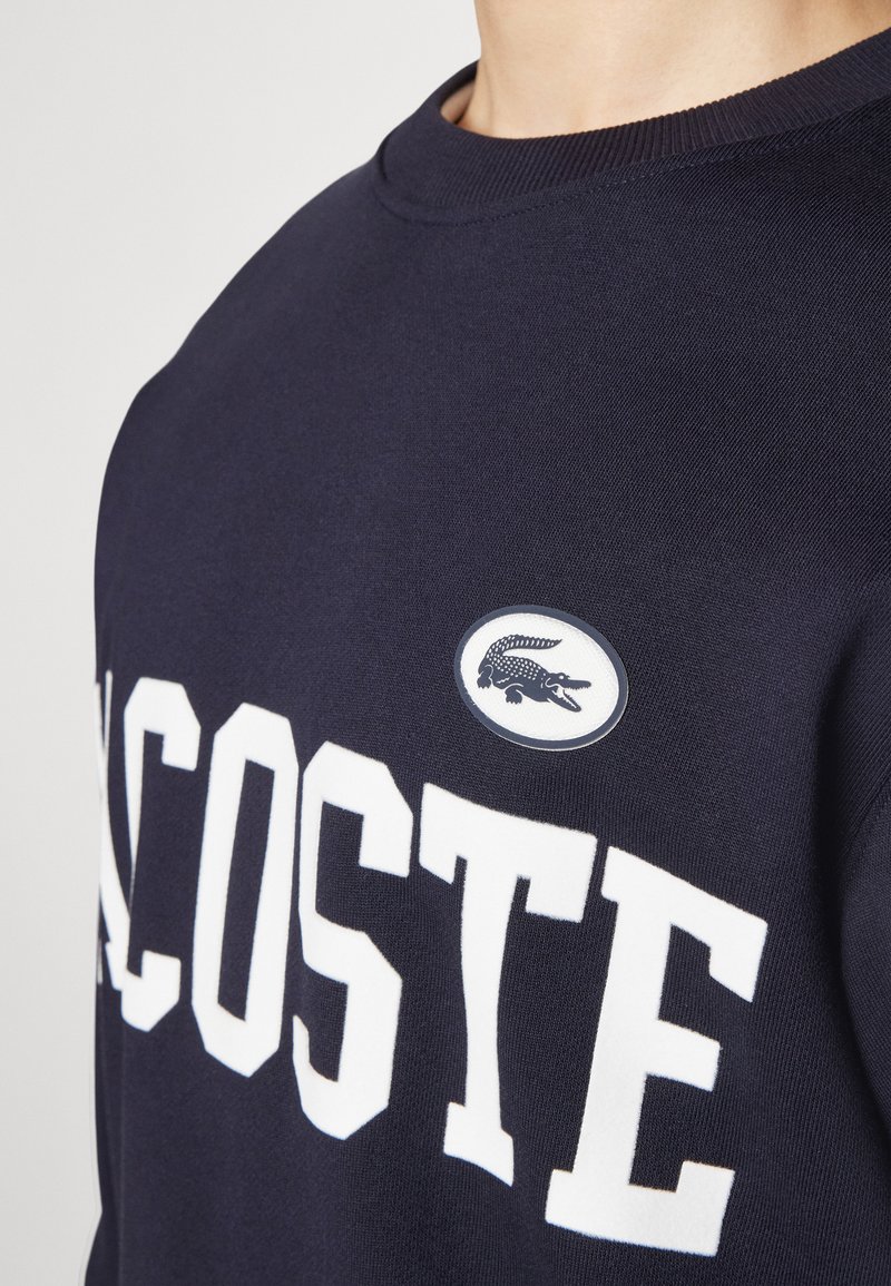 Sudadera navy con letras blancas "LACOSTE", cuello redondo acanalado y logotipo ovalado con un cocodrilo. La tela tiene una textura suave.