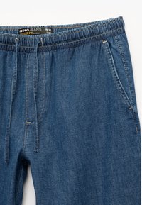 Pantalones jogger en azul oscuro hechos de denim, con cinturilla elástica y cordón, bolsillos laterales y detalles de costura visibles.