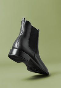 Stivale Chelsea in pelle nera con pannelli laterali elastici e linguetta, inclinato per mostrare la suola e le cuciture su uno sfondo verde chiaro uniforme.