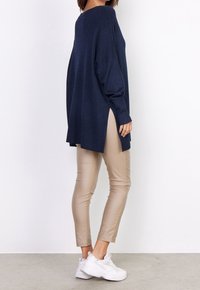 Marineblauer übergroßer Strickpullover mit seitlichen Schlitzen, kombiniert mit beigefarbenen, eng anliegenden Hosen und weißen Sportschuhen, gezeigt in der Seitenansicht.