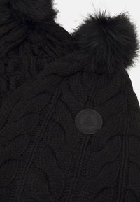 Bonnet en tricot noir avec des motifs en câble, deux pompons noirs duveteux et un patch en caoutchouc rond texturé de la marque sur le côté.