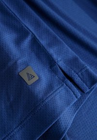 Niebieska tkanina sportowa o teksturowanym, przewiewnym wzorze. Zawiera małą czarną naszywkę z logo oraz szczegóły szwów, świadczące o wysokowydajnym designie.