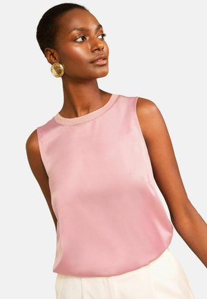 Nő, aki világos rózsaszín ujjatlan selyem blúzt és nagy, kerek arany fülbevalókat visel, balra néz sima háttér előtt.