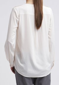 Chemisier boutonné en tissu doux couleur crème avec une coupe ample, manches longues et encolure ronde. Comprend un empiècement au dos et un léger éclat.