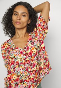 Blusa floral com decote em V, mangas curtas e bainha com babados. Apresenta padrões vibrantes em vermelho, amarelo e verde sobre um fundo claro.