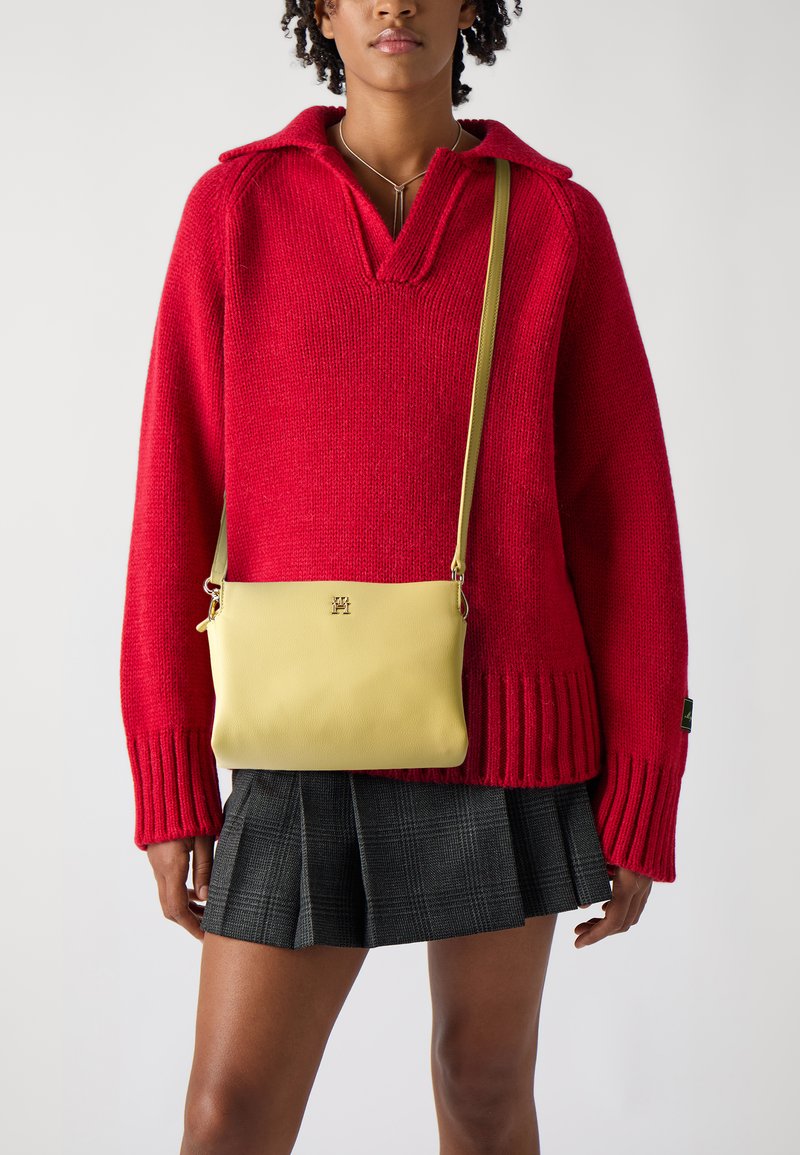 Žlutá kožená crossbody taška se zlatým kováním, obdélníkového tvaru a nastavitelným popruhem, nošená přes červený pletený svetr a kostkovanou sukni.