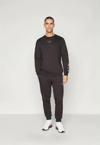 Hombre de pie contra un fondo liso, vistiendo un sudadera de manga larga negra y joggers a juego, con zapatillas blancas.