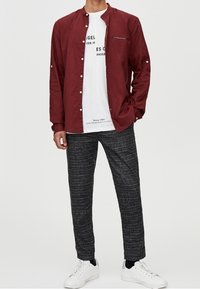 Chemise bordeaux à boutons avec col montant, associée à un t-shirt blanc à motif et un pantalon gris à motifs. Des baskets blanches complètent la tenue.