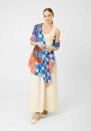 Femme portant une robe crème sans manches et un châle coloré bleu, orange et rose à motifs posé sur les épaules sur fond blanc.