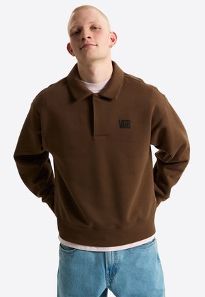Junger Mann mit heller Haut und kurzem blondem Haar, der einen braunen Vans-Pullover und hellblaue Jeans trägt, steht mit den Händen hinter dem Rücken.