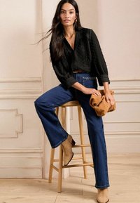 Blusa nera con texture e maniche a tre quarti, jeans a campana blu, stivali ankle beige con tacco, e una clutch marrone appoggiata su uno sgabello di legno.