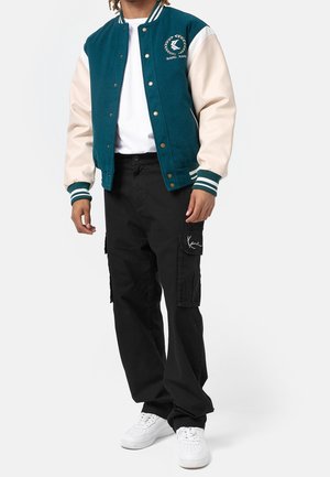Personne portant une veste universitaire bleu sarcelle et crème, un t-shirt blanc, un pantalon cargo noir avec logo sur la poche, et des baskets blanches, debout devant un fond blanc.