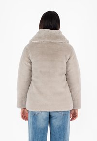 Giacca in finta pelliccia grigia chiara con ampio colletto, maniche lunghe e una consistenza morbida, indossata sopra jeans di denim blu. Vista posteriore mostrata.