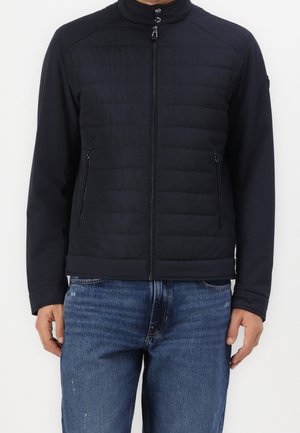 Mand iført sort quiltet jakke med lynlåslommer og mørkeblå jeans, stående mod en hvid baggrund.