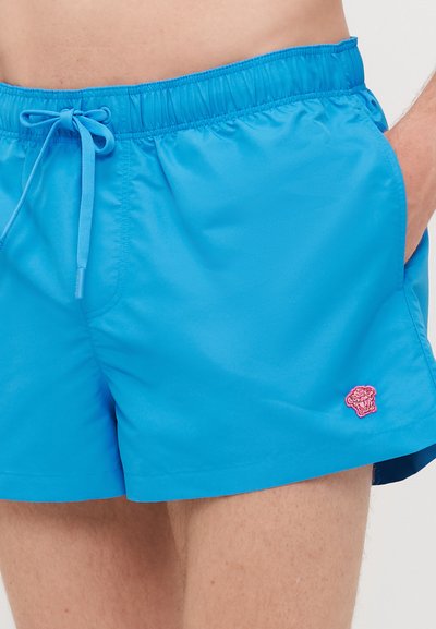 Shorts de bain bleus avec une taille élastique et un cordon de serrage, ornés d'un emblème rose brodé sur le côté gauche. Tissu lisse et léger.