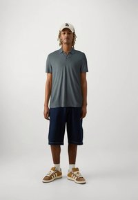 Jeune homme portant un polo gris à manches courtes, un short noir jusqu'aux genoux, des chaussettes blanches, des baskets beiges et une casquette blanche LA, se tenant devant un fond uni.