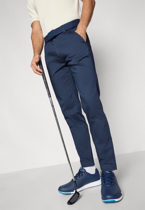 Persona que lleva pantalones y zapatos azul marino, sostiene un palo de golf con una mano y tiene la otra en el bolsillo, de pie sobre un suelo gris frente a un fondo liso.