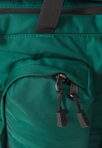 Mochila verde em nylon com tecido texturizado, apresentando um bolso frontal e zíperes duplos com cordões pretos e detalhes de marca.